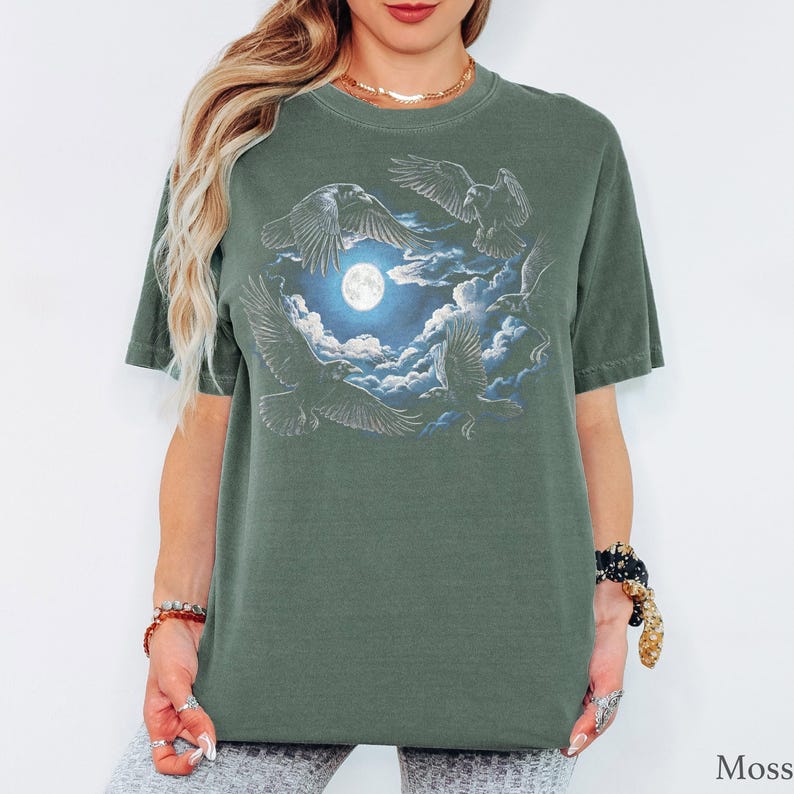 May include: A moss green short-sleeve t-shirt featuring a graphic print of four black ravens flying around a bright full moon surrounded by white clouds. The design is centered on the chest of the shirt, which has a crew neckline and a relaxed fit.