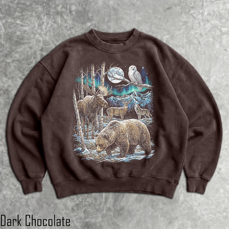 Pu&ograve; includere: Felpa color cioccolato fondente con una scena di foresta con un orso, un alce, cervi e un lupo sotto l'aurora boreale. Un gufo &egrave; appollaiato su un ramo vicino alla luna piena. Il testo "Dark Chocolate" &egrave; in basso.