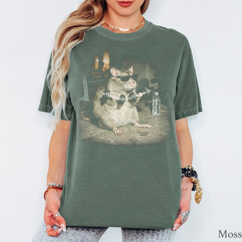 May include: A moss green short-sleeve t-shirt featuring a graphic print of a brown rat wearing black sunglasses and lifting two small dumbbells. The rat is positioned in the center of the shirt, with a water bottle visible in the background.