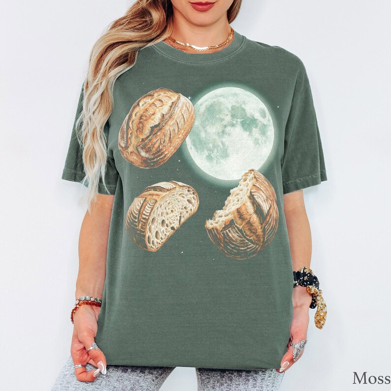 May include: A moss green short-sleeve t-shirt featuring a graphic print of a full moon and three loaves of sourdough bread. The design includes one whole loaf, one sliced loaf, and one partially eaten loaf arranged around the glowing white moon.
