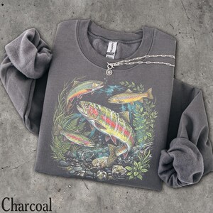 May include: A charcoal gray long-sleeve sweatshirt featuring a graphic print of three trout swimming among aquatic plants and rocks. The sweatshirt has a crew neckline and a Gildan brand label at the inner collar. A silver chain necklace is visible.