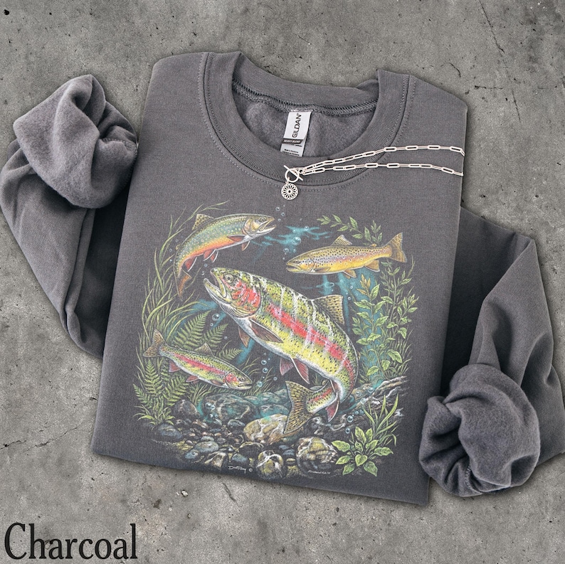 May include: A charcoal gray long-sleeve sweatshirt featuring a graphic print of three trout swimming among aquatic plants and rocks. The sweatshirt has a crew neckline and a Gildan brand label at the inner collar. A silver chain necklace is visible.