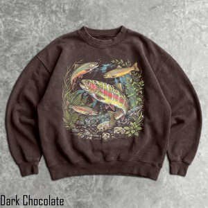 May include: A dark chocolate brown crewneck sweatshirt featuring a detailed graphic print of three rainbow trout swimming among green aquatic plants and rocks. The illustration displays the fish with vibrant scales and distinct markings.