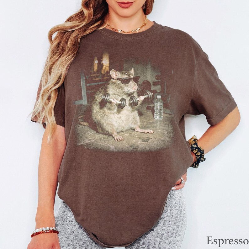 May include: An espresso-colored short-sleeve t-shirt featuring a graphic print of a rat wearing black sunglasses and lifting two silver dumbbells in a gym setting, with a clear water bottle placed on the floor next to the rat.