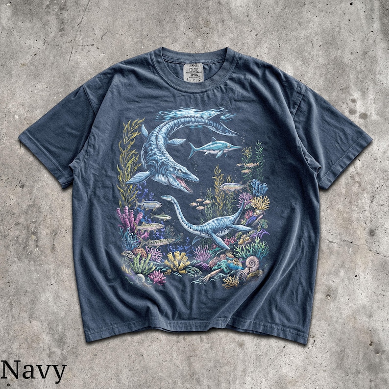 Op de afbeelding: Marineblauw T-shirt met een gedetailleerde illustratie van zeereptielen en vissen in een onderwaterlandschap. Het ontwerp bevat een grote plesiosaurus en andere zeedieren te midden van kleurrijk koraal en zeewier. Het woord "Navy" staat onderaan gedrukt.