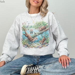 Op de afbeelding: Een lichtgrijs sweatshirt met ronde hals en een kleurrijke grafische print van otters die in een rivier zwemmen tussen waterplanten, rotsen en een reiger. Het ontwerp toont een gedetailleerd tafereel van dieren in hun natuurlijke habitat.