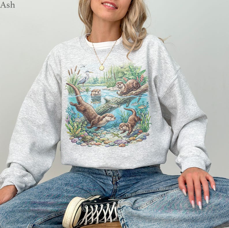 Op de afbeelding: Een lichtgrijs sweatshirt met ronde hals en een kleurrijke grafische print van otters die in een rivier zwemmen tussen waterplanten, rotsen en een reiger. Het ontwerp toont een gedetailleerd tafereel van dieren in hun natuurlijke habitat.