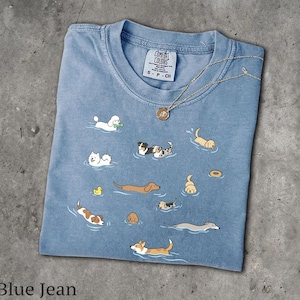 Può includere: Una t-shirt color blu jeans con una stampa grafica di varie razze canine che nuotano nell'acqua. La maglietta presenta uno scollo rotondo e un'etichetta del marchio Comfort Colors all'interno del colletto. Una sottile catenina dorata poggia sul tessuto.