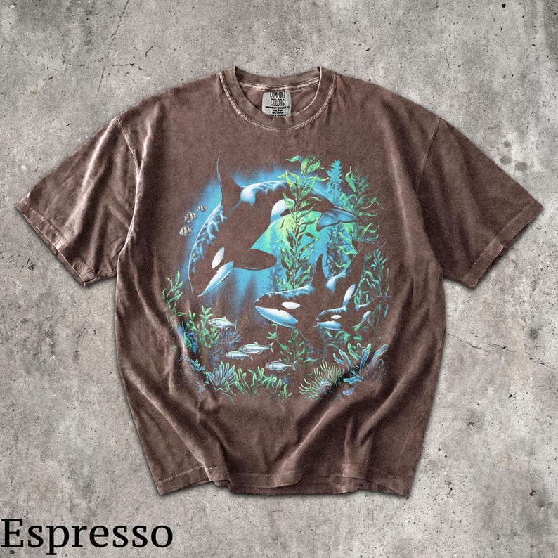 May include: A brown short-sleeve t-shirt featuring a graphic print of two orcas swimming among green seaweed and small fish. The design is set against a blue circular background. The shirt has a crew neckline and a Comfort Colors brand label.