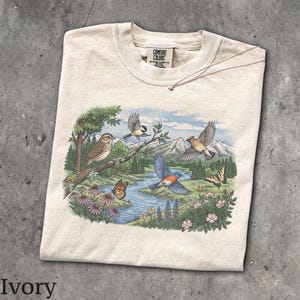 May include: An ivory t-shirt featuring a graphic print of a nature scene with birds, butterflies, a river, and mountains. The Comfort Colors brand label is visible at the inner neckline. The illustration includes a bluebird, a chickadee, and various wildflowers in a landscape setting.