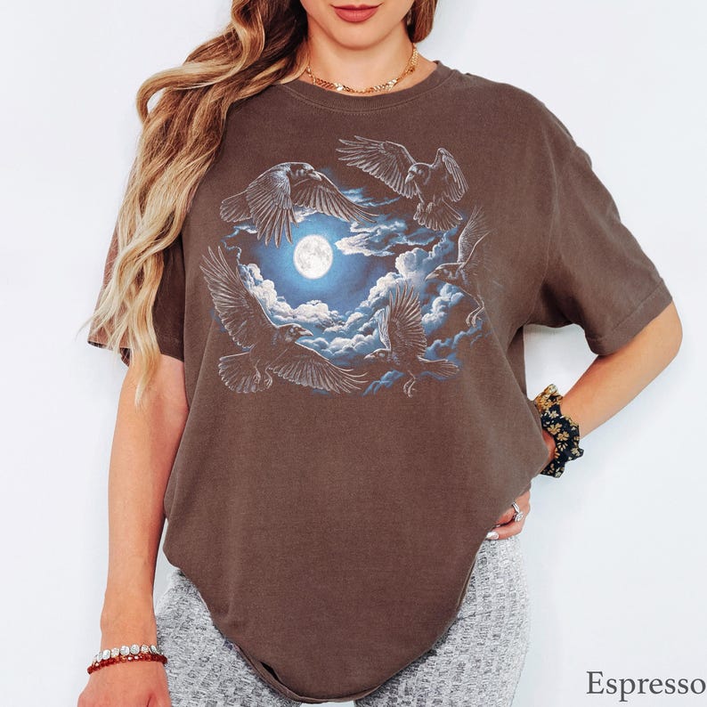 May include: An espresso brown short-sleeve t-shirt featuring a graphic print of four black ravens flying in a circular formation around a bright white full moon set against a backdrop of blue and white clouds. The word Espresso is printed below.
