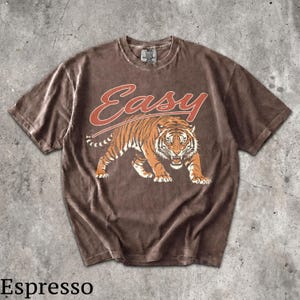 May include: A brown short-sleeve t-shirt featuring a graphic of a snarling tiger in orange and black tones. Above the tiger, the word "Easy" is printed in a bold, orange script font. The shirt has a crew neckline and a faded, washed-out appearance.
