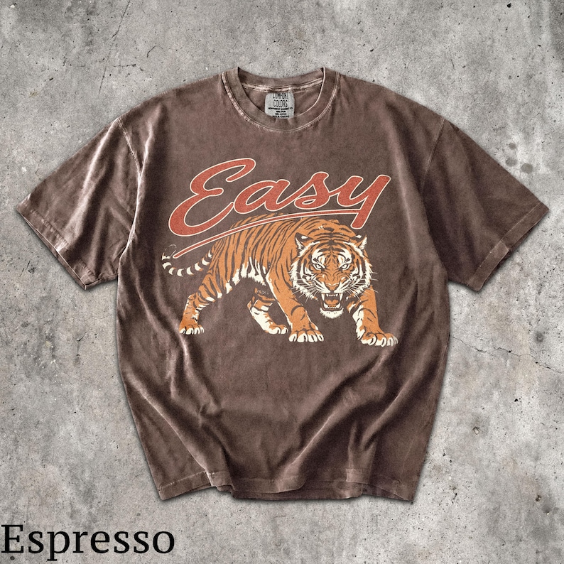 May include: A brown short-sleeve t-shirt featuring a graphic of a snarling tiger in orange and black tones. Above the tiger, the word "Easy" is printed in a bold, orange script font. The shirt has a crew neckline and a faded, washed-out appearance.