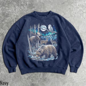 Pu&ograve; includere: Felpa blu navy con un'illustrazione dettagliata della fauna selvatica in una foresta innevata. Il design include un orso, un alce, cervi e un lupo sotto la luna piena e l'aurora boreale. Un gufo &egrave; appollaiato su un ramo. La parola "Navy" &egrave; in basso.