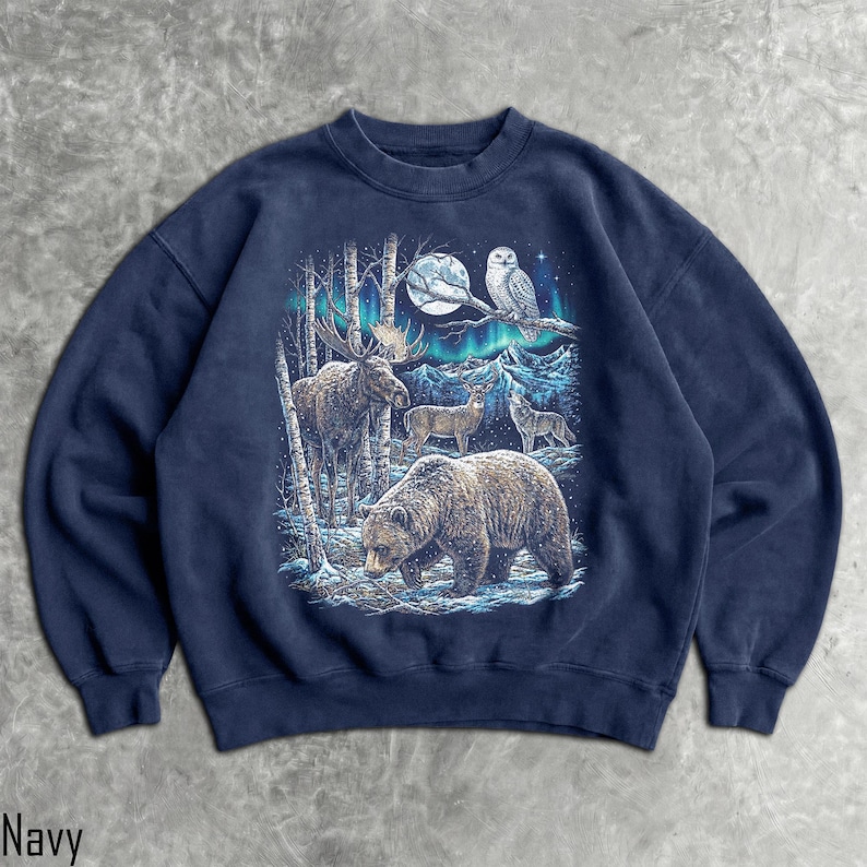 Pu&ograve; includere: Felpa blu navy con un'illustrazione dettagliata della fauna selvatica in una foresta innevata. Il design include un orso, un alce, cervi e un lupo sotto la luna piena e l'aurora boreale. Un gufo &egrave; appollaiato su un ramo. La parola "Navy" &egrave; in basso.