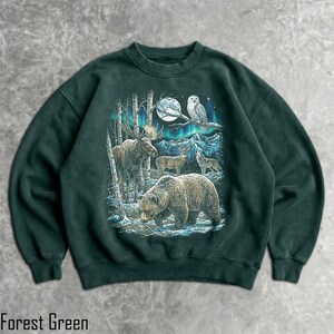 Pu&ograve; includere: Felpa verde foresta con un disegno di fauna selvatica raffigurante un orso, un alce, un cervo, un lupo e un gufo sotto la luna piena e l'aurora boreale. Il testo "Forest Green" &egrave; stampato in basso nell'immagine.