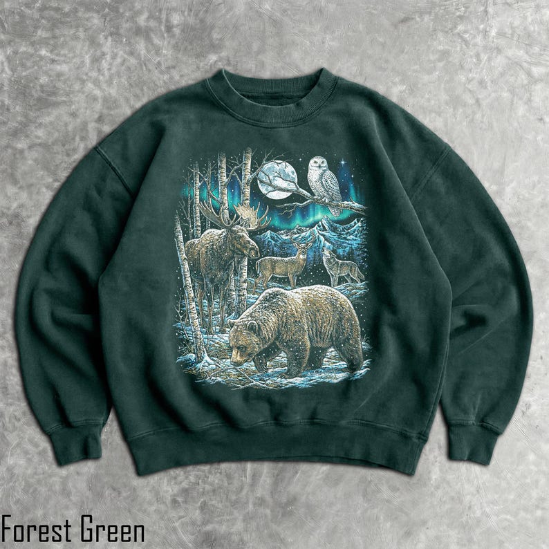 Pu&ograve; includere: Felpa verde foresta con un disegno di fauna selvatica raffigurante un orso, un alce, un cervo, un lupo e un gufo sotto la luna piena e l'aurora boreale. Il testo "Forest Green" &egrave; stampato in basso nell'immagine.