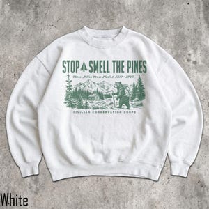 Pode incluir: Uma camisola branca de gola redonda com uma impress&atilde;o gr&aacute;fica verde-escura. O texto diz STOP AND SMELL THE PINES, Three Billion Trees Planted 1933-1942 e CIVILIAN CONSERVATION CORPS. A ilustra&ccedil;&atilde;o mostra um urso com mochila numa floresta.