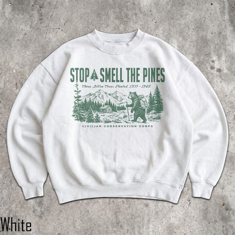 Pode incluir: Uma camisola branca de gola redonda com uma impress&atilde;o gr&aacute;fica verde-escura. O texto diz STOP AND SMELL THE PINES, Three Billion Trees Planted 1933-1942 e CIVILIAN CONSERVATION CORPS. A ilustra&ccedil;&atilde;o mostra um urso com mochila numa floresta.
