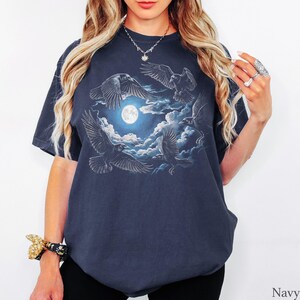 May include: A navy blue short-sleeve t-shirt featuring a graphic print of four black ravens flying around a bright white full moon surrounded by dark blue clouds. The design is centered on the chest of the garment, which has a crew neckline.