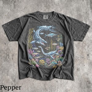 Op de afbeelding: Donkergrijs t-shirt met een gedetailleerde illustratie van prehistorische zeereptielen, waaronder een mosasaurus en plesiosaurus, zwemmend tussen kleurrijk koraal en vissen. Het woord "Pepper" staat onderaan de afbeelding.