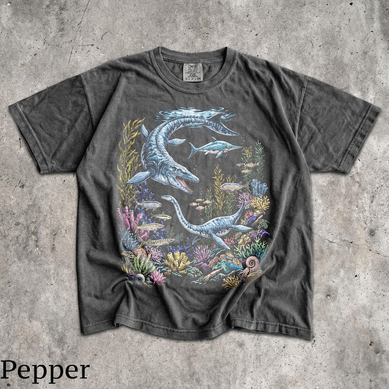 Op de afbeelding: Donkergrijs t-shirt met een gedetailleerde illustratie van prehistorische zeereptielen, waaronder een mosasaurus en plesiosaurus, zwemmend tussen kleurrijk koraal en vissen. Het woord "Pepper" staat onderaan de afbeelding.