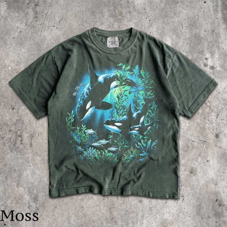 May include: A moss green short-sleeve t-shirt featuring a graphic print of two orcas swimming among underwater kelp and small fish. The design includes a blue light effect behind the whales. The shirt has a crew neckline and a Comfort Colors label.