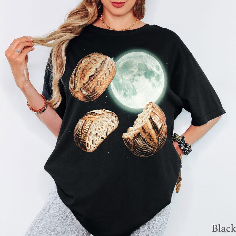 May include: A black short-sleeve t-shirt featuring a graphic print of a large, glowing full moon surrounded by three loaves of crusty, golden-brown sourdough bread. The design includes small white star-like dots scattered across the dark background.