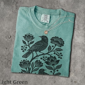 May include: A light green t-shirt featuring a black graphic print of a bird perched on a branch surrounded by flowers and berries. The shirt has a crew neckline with a Comfort Colors label showing size S-CH and a thin silver chain necklace resting on the collar.