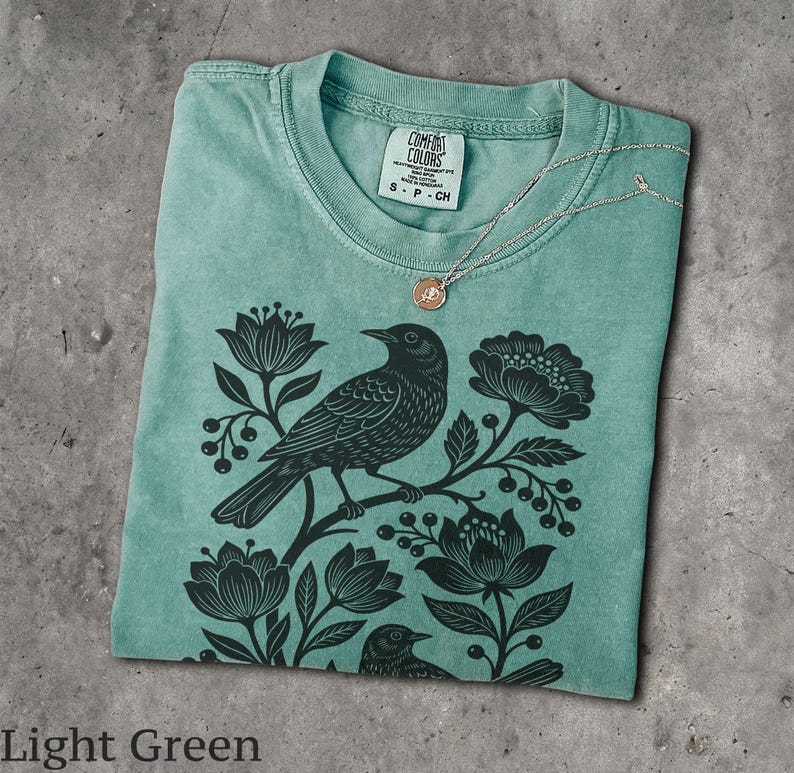 May include: A light green t-shirt featuring a black graphic print of a bird perched on a branch surrounded by flowers and berries. The shirt has a crew neckline with a Comfort Colors label showing size S-CH and a thin silver chain necklace resting on the collar.