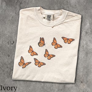May include: An ivory short-sleeve t-shirt featuring a graphic print of six orange and black monarch butterflies arranged in a diagonal pattern. The inner neck label displays the text Comfort Colors, 100% cotton, and made in Honduras.