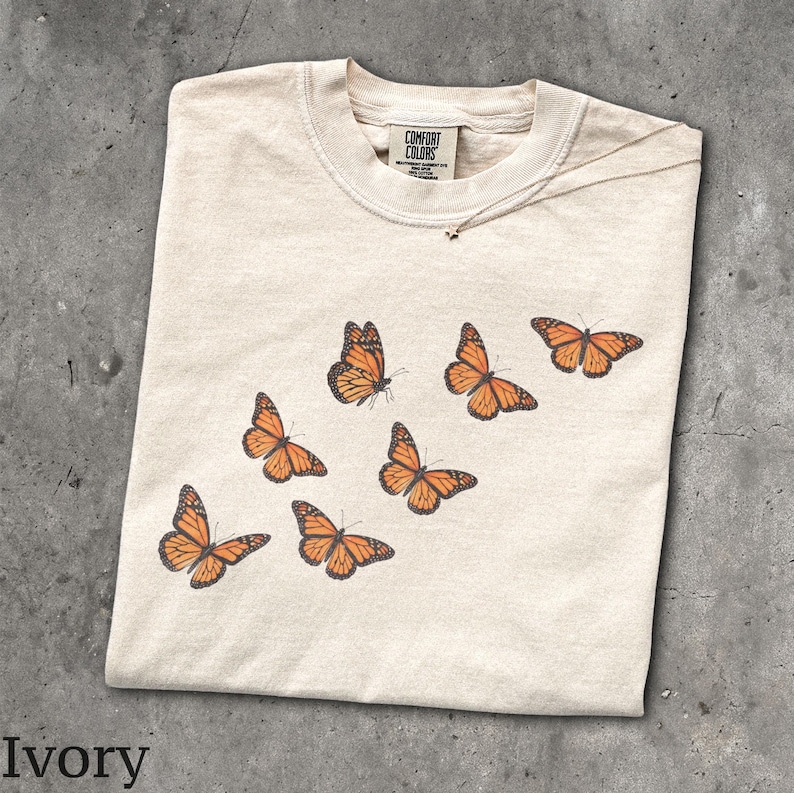 May include: An ivory short-sleeve t-shirt featuring a graphic print of six orange and black monarch butterflies arranged in a diagonal pattern. The inner neck label displays the text Comfort Colors, 100% cotton, and made in Honduras.