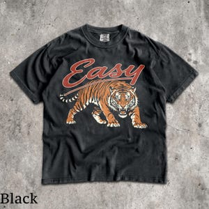 May include: A black short-sleeve t-shirt featuring a graphic of a prowling tiger in orange and black tones. Above the tiger, the word "Easy" is printed in a stylized red script font. The shirt has a crew neckline and a standard fit design.
