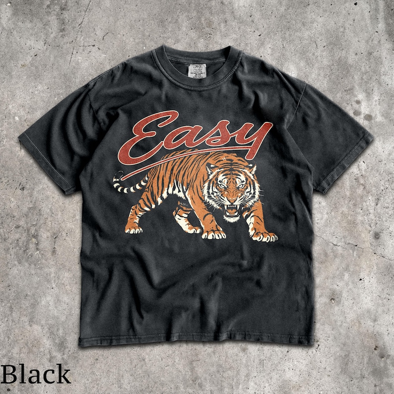 May include: A black short-sleeve t-shirt featuring a graphic of a prowling tiger in orange and black tones. Above the tiger, the word "Easy" is printed in a stylized red script font. The shirt has a crew neckline and a standard fit design.