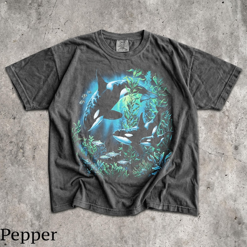 May include: A charcoal gray short-sleeve t-shirt featuring a graphic print of two orcas swimming among green seaweed and small fish. The design is centered on the chest, and the shirt has a crew neckline with a visible brand tag at the collar.