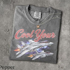 Może przedstawiać: Ciemnoszary t-shirt z krótkim rękawem, okrągłym dekoltem i metką Comfort Colors. Grafika przedstawia dwa odrzutowce w locie z pomarańczowymi płomieniami z silników oraz czerwonym napisem Cool Your. Na koszulce leży cienki, złoty łańcuszek.