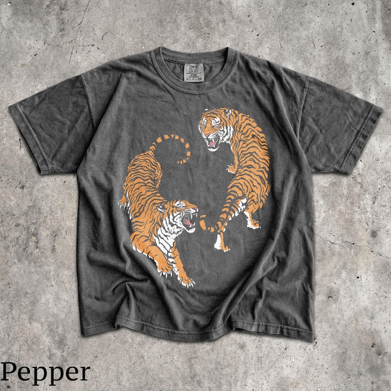 May include: A charcoal gray short-sleeve t-shirt featuring a graphic of two orange and black tigers in a dynamic pose. The tigers are positioned in a circular arrangement with their mouths open. The shirt has a crew neckline and a faded, washed-out appearance.
