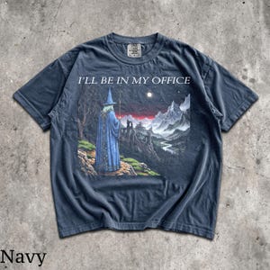 Op de afbeelding: Een marineblauw t-shirt met korte mouwen en een grafische print van een tovenaar in een blauwe mantel met een staf, voor een berglandschap met een kasteel en de tekst I'LL BE IN MY OFFICE in het wit boven de illustratie.
