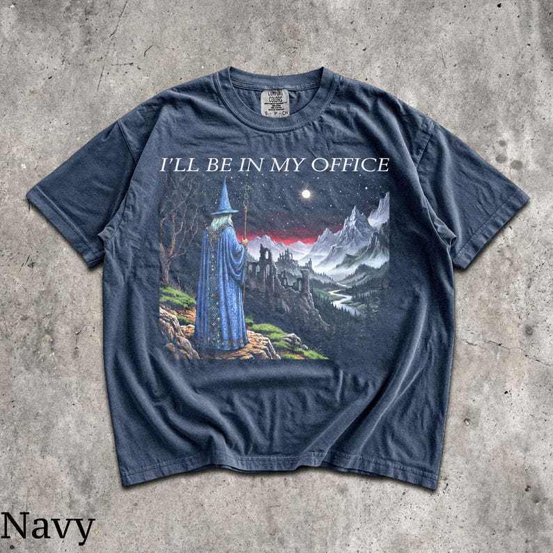 Op de afbeelding: Een marineblauw t-shirt met korte mouwen en een grafische print van een tovenaar in een blauwe mantel met een staf, voor een berglandschap met een kasteel en de tekst I'LL BE IN MY OFFICE in het wit boven de illustratie.