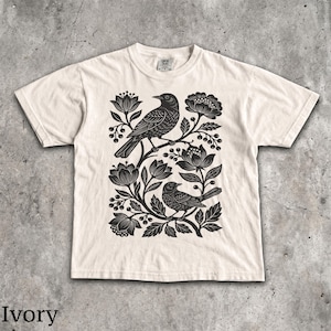 May include: An ivory short-sleeve t-shirt featuring a black graphic print of two birds perched among stylized flowers, leaves, and berries. The design is centered on the chest, and the shirt has a crew neckline and a standard fit.