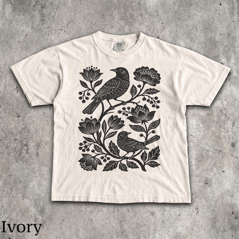 May include: An ivory short-sleeve t-shirt featuring a black graphic print of two birds perched among stylized flowers, leaves, and berries. The design is centered on the chest, and the shirt has a crew neckline and a standard fit.