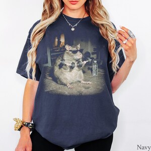 May include: A navy blue t-shirt featuring a graphic print of a brown rat wearing black sunglasses and lifting two small silver dumbbells. The rat is positioned in a gym setting with a clear plastic water bottle visible on the floor nearby.