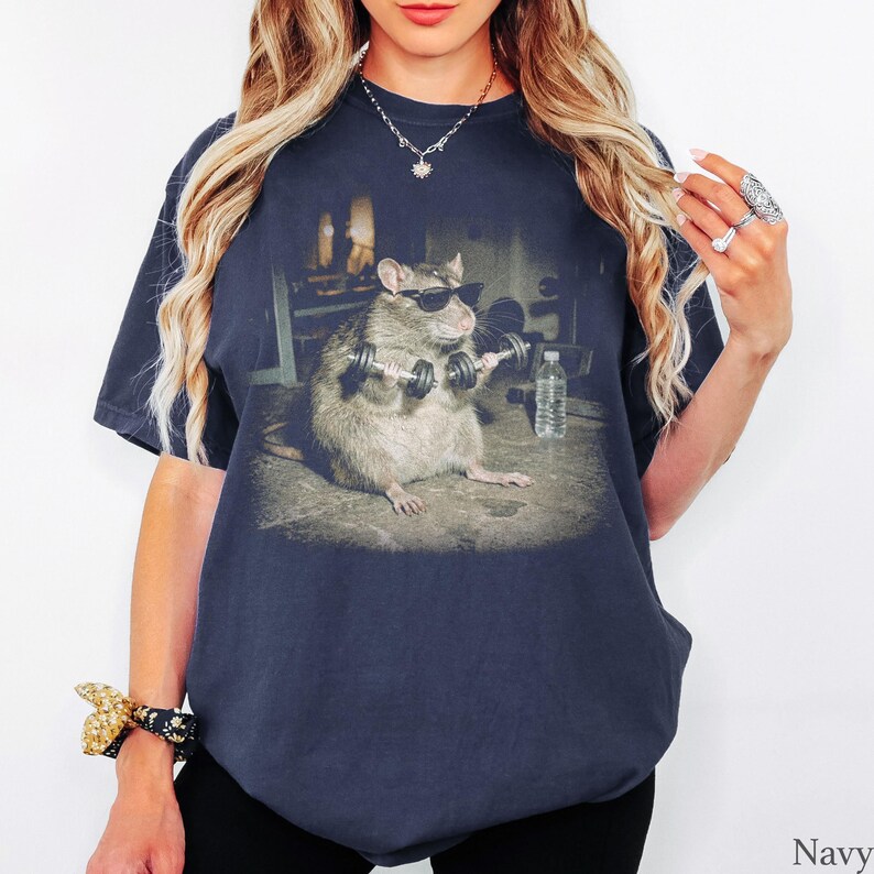 May include: A navy blue t-shirt featuring a graphic print of a brown rat wearing black sunglasses and lifting two small silver dumbbells. The rat is positioned in a gym setting with a clear plastic water bottle visible on the floor nearby.