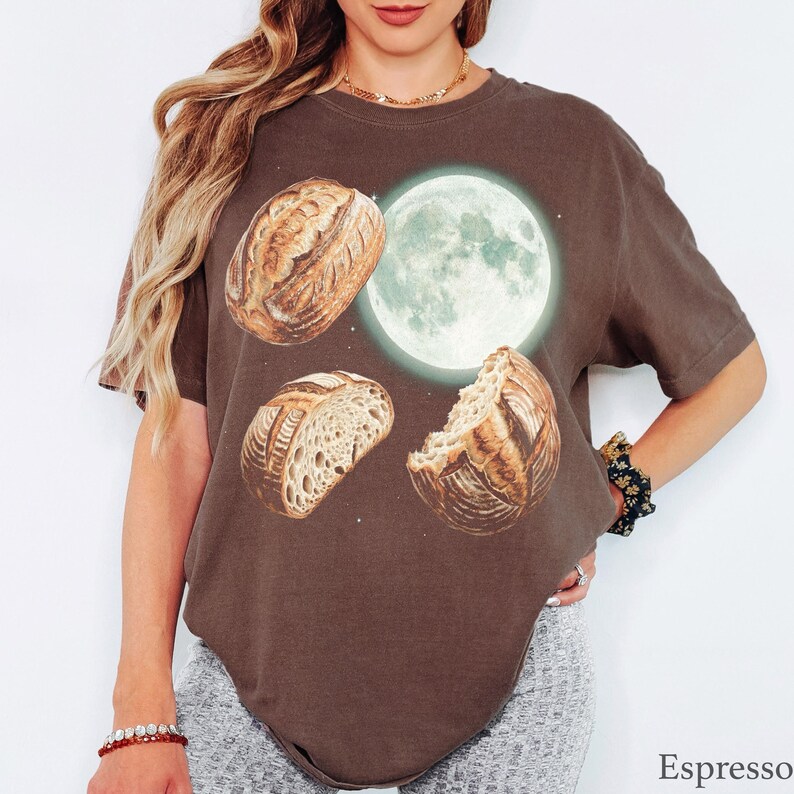 May include: A dark brown short-sleeve t-shirt featuring a graphic print of three sourdough bread loaves arranged around a glowing full moon. The text "Espresso" is printed in the bottom right corner of the image.
