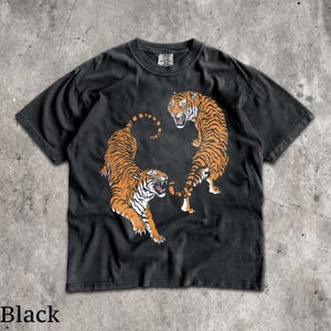 May include: A black short-sleeve t-shirt featuring a graphic print of two orange and black tigers in a dynamic pose. The tigers are positioned to face each other with mouths open, displaying white teeth. The shirt has a crew neckline and a tag.