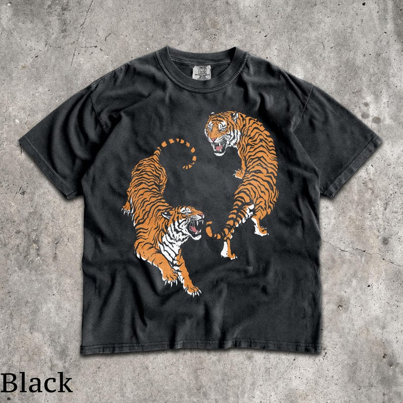 May include: A black short-sleeve t-shirt featuring a graphic print of two orange and black tigers in a dynamic pose. The tigers are positioned to face each other with mouths open, displaying white teeth. The shirt has a crew neckline and a tag.