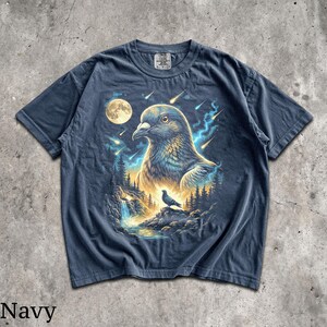 May include: A navy blue short-sleeve t-shirt featuring a graphic print of a large pigeon head in the sky above a smaller pigeon perched on a rock near a waterfall, surrounded by a forest, a full moon, and streaks of light in the night sky.