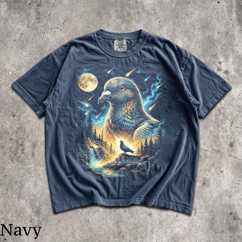 May include: A navy blue short-sleeve t-shirt featuring a graphic print of a large pigeon head in the sky above a smaller pigeon perched on a rock near a waterfall, surrounded by a forest, a full moon, and streaks of light in the night sky.