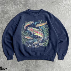 May include: A navy blue crewneck sweatshirt featuring a detailed graphic print of three rainbow trout swimming among aquatic plants and rocks. The central fish is large and colorful, while two smaller fish are positioned above and below it.