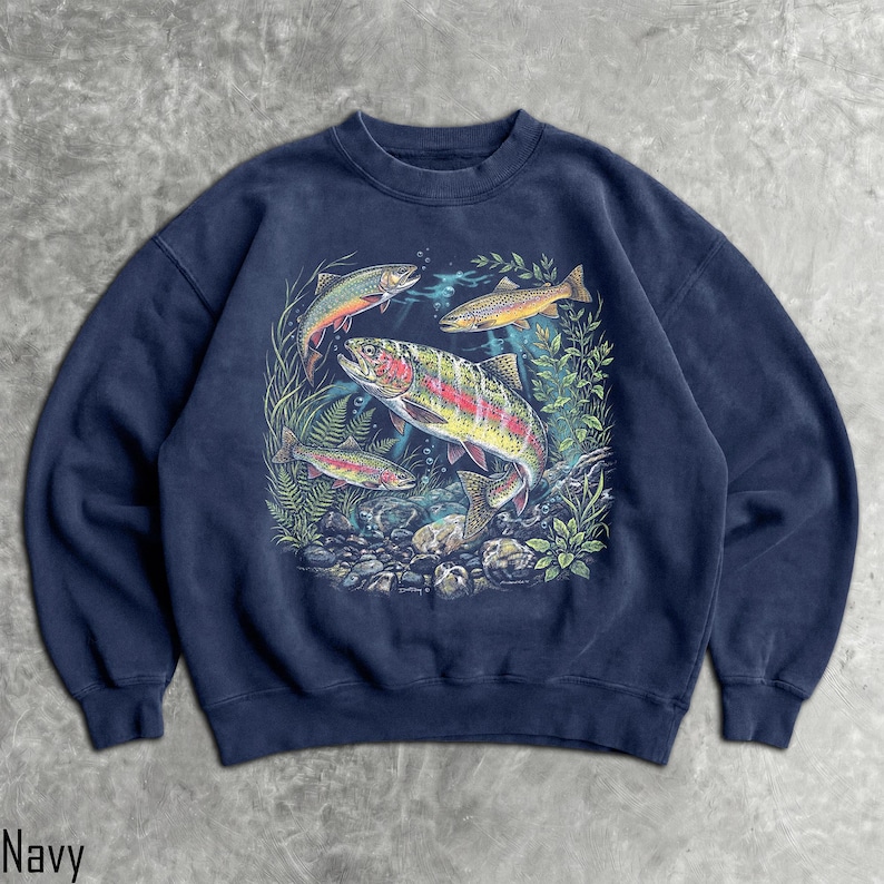 May include: A navy blue crewneck sweatshirt featuring a detailed graphic print of three rainbow trout swimming among aquatic plants and rocks. The central fish is large and colorful, while two smaller fish are positioned above and below it.
